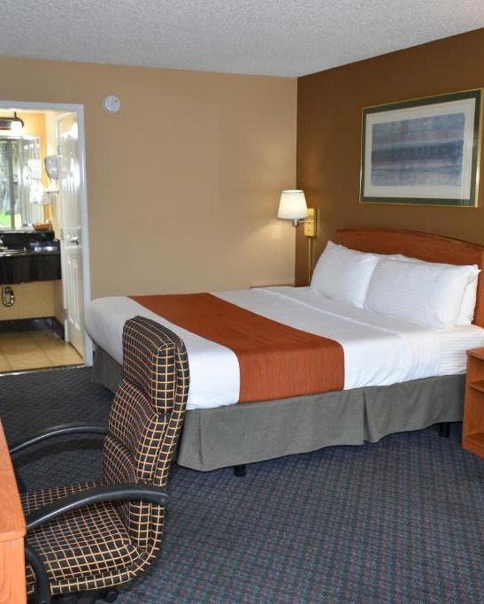 A hotel room with a bed, two nightstands, lamps, a desk and chair, an armchair, and an adjacent bathroom area, neat and functional.