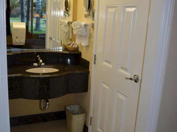 A bathroom with a dark granite sink counter, a oval mirror, towels on a rack, a white door, and a small trash can by the sink, with tiled floor.