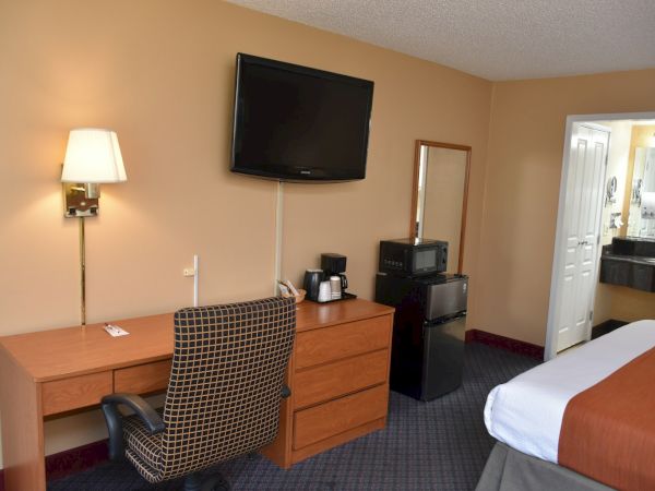 A hotel room with a desk and chair, wall-mounted TV, mini fridge, microwave, coffee maker, mirror, and a bed with brown bedding; doorway to a bathroom.