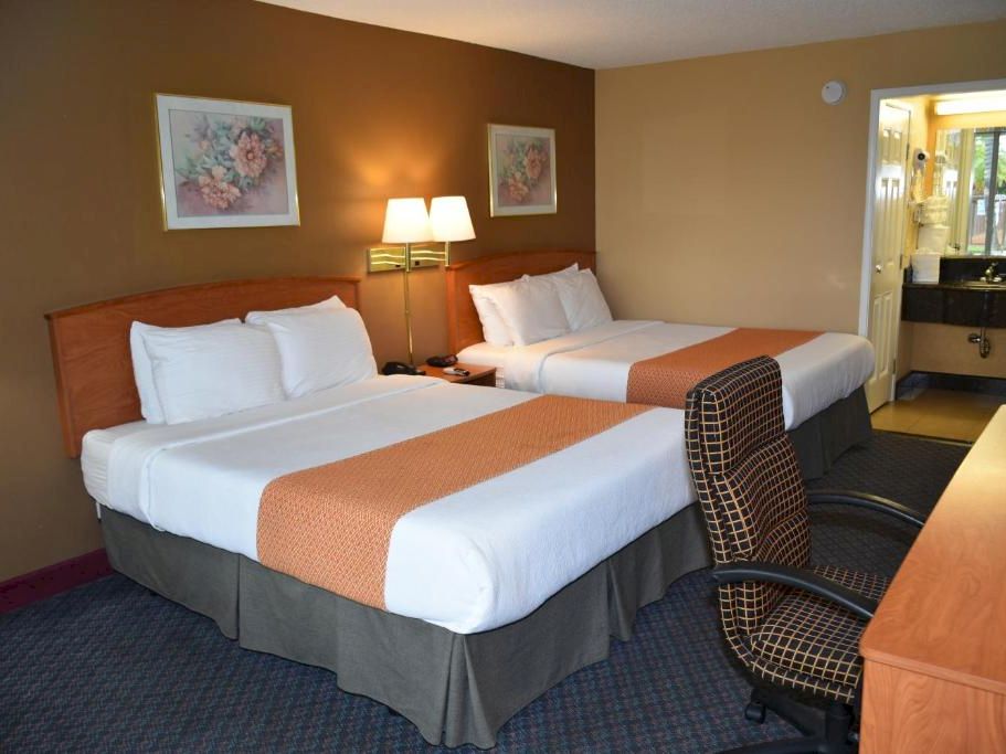 Two double beds with white linens and orange bed runners, a desk and chair, lamps, artwork, and a view into a small bathroom.