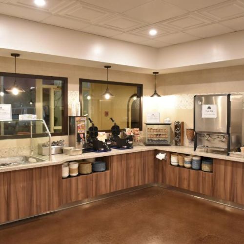 A cozy cafe counter with warm wood cabinets, a coffee station, self-serve condiments, and hanging pendant lights by a large window.
