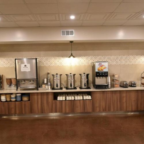 A long breakfast buffet setup with coffee dispensers, drink machines, warm lights, and assorted food stations along a wooden counter, empty.
