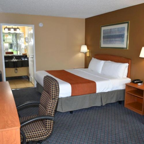 A hotel room with a bed, nightstands, lamps, a desk and chair, a small sofa, and an open bathroom doorway in the back, neat and simple.