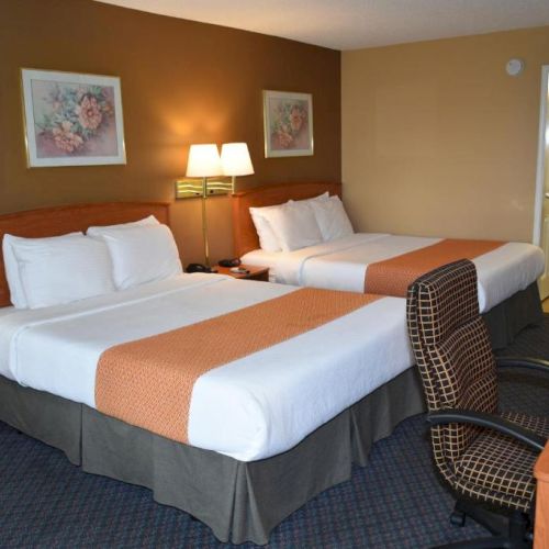 Two double beds with white linens and orange accents, a desk chair, nightstand with lamps, and a doorway to a bathroom.