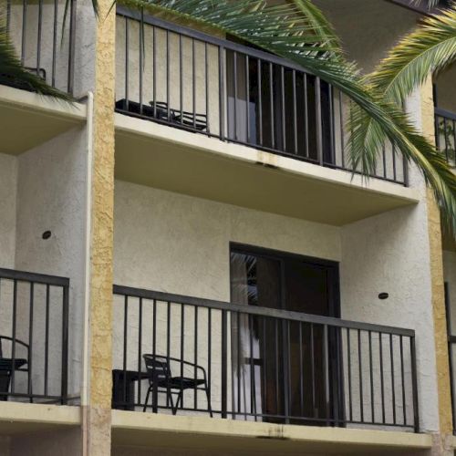 A row of small balconies on a beige apartment building, with black railings, chairs, a table, palm trees, and tropical plants.