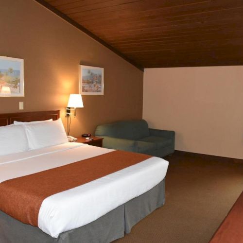 A hotel room with a large bed, white linens and brown bed runner, a sofa, desk, lamps, framed pictures, and a window with curtains, cozy and tidy.