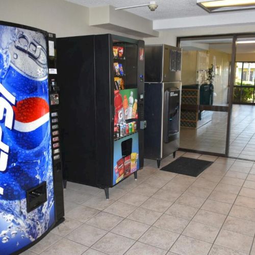 A hallway with a Pepsi vending fridge on the left, open snack shelves, and glass doors leading to another room, bright and tiled.