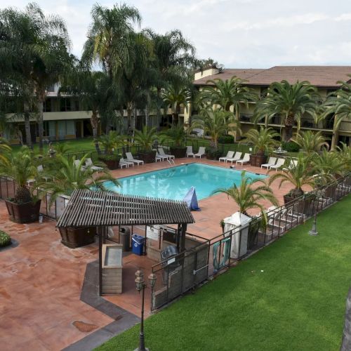 An outdoor resort pool area with a rectangular pool, palm trees, lounge chairs, a shaded gazebo, brick patio, and green lawns surrounding a multi-building complex.