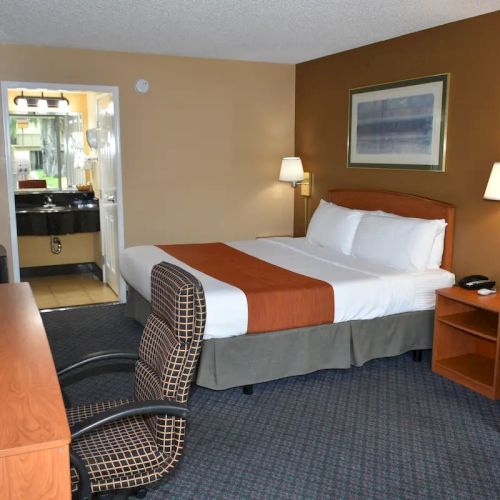 A hotel room with a bed, nightstands, lamps, desk, chair, and a doorway to the bathroom area, in warm tones.