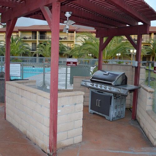 A fenced pool area with a covered brick barbecue station, a gas grill, and surrounding palm trees at a resort.