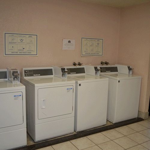 A row of four top-load washers in a laundry room, with framed certificates on the wall and a Pepsi vending machine on the right.