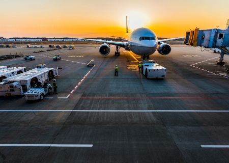 A large airplane at the gate during sunset, with ground support vehicles nearby and a jet bridge in the background, on the tarmac.