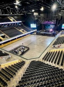 A large indoor arena set up for an event with a central floor stage, seating surrounding on all sides, two large screens, and lighting rig. Top it at 140 characters, always ending the sentence.