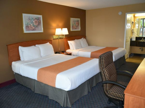 Two double beds with white linens and orange runners, a nightstand lamp, framed art, and a small desk facing a doorway to a bathroom.