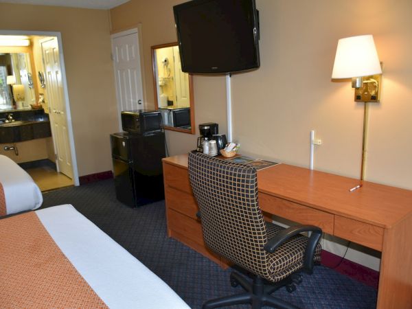 A hotel room with a desk, chair, wall-mounted TV, mini-fridge, coffee maker, lamp, and a bed partly visible; doorway to a bathroom in the back.