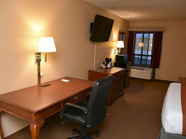 A hotel room with a wooden desk, rolling chair, wall lamp, TV on the wall, a dresser with a coffee maker, window with curtains, and a bed.