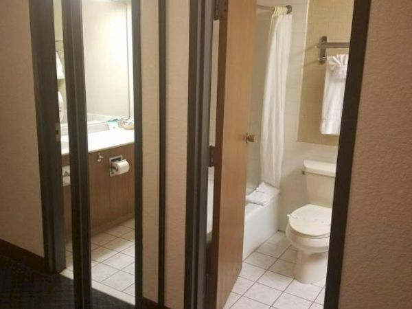 A small hotel bathroom visible through open doors: sink, mirror, toilet, and shower with a white curtain; tile floor, neutral tones.