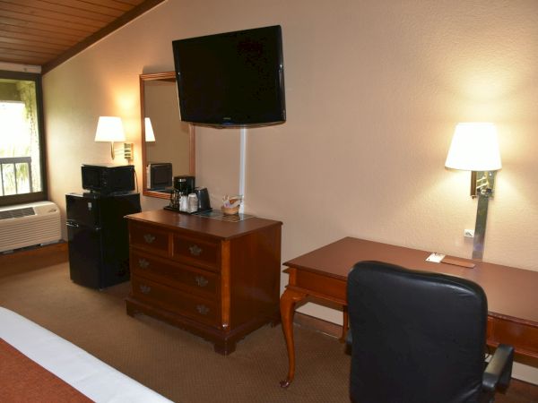 A hotel room with a desk, chair, dresser, wall-mounted TV, small table lamps, and a window seat near the bed, plus a mini-fridge area and a TV on the dresser.