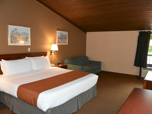 A hotel room with a large bed, white linens and brown throw, a small sofa, bedside lamps, framed pictures, and a wooden ceiling, simple and tidy.