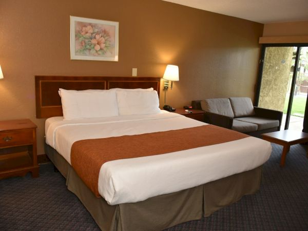 A hotel room with a large bed, white linens, brown bed runner, two nightstands with lamps, a sofa, coffee table, and sliding door to a patio.