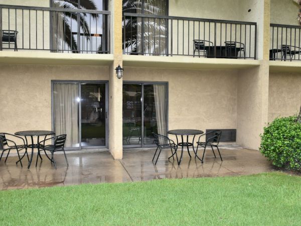 Patio with outdoor tables and chairs on a wet concrete walkway, two sliding doors to a beige building, balconies above, a green lawn in the foreground.