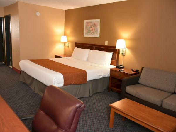 A hotel room with a king bed, two nightstands, a sofa, a coffee table, lamps, and a small desk area, in warm brown-beige tones.
