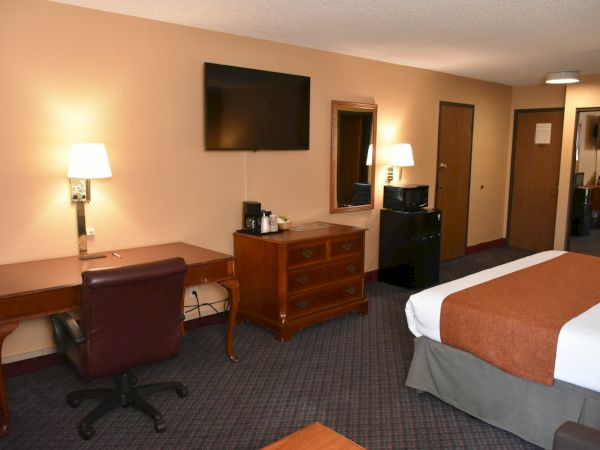 A hotel room with a desk, chair, dresser, wall-mounted TV, mirror, two lamps, a coffee/tea station, and a bed with brown and white bedding.