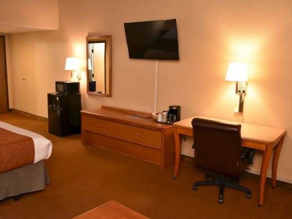 A hotel room with a bed, two lamps, a dresser, wall-mounted TV, a desk and chair, and a few electronics on the dresser.