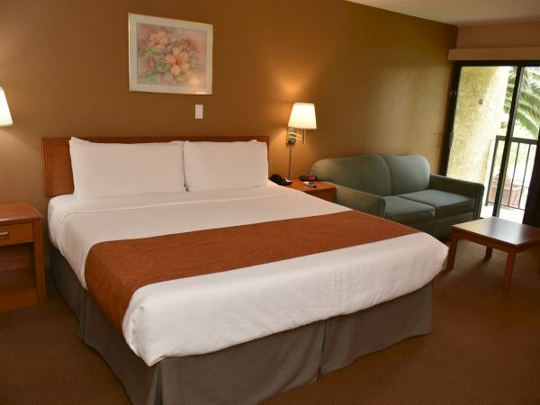 A hotel room with a large bed, two nightstands, lamps, a sofa, coffee table, artwork, and a balcony door.