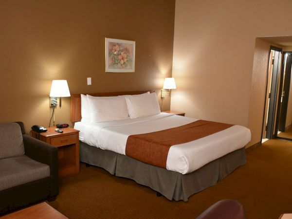 A hotel room with a king bed, white linens, brown bed scarf, two bedside lamps, a small sofa, and a nightstand; doorway to a corridor.