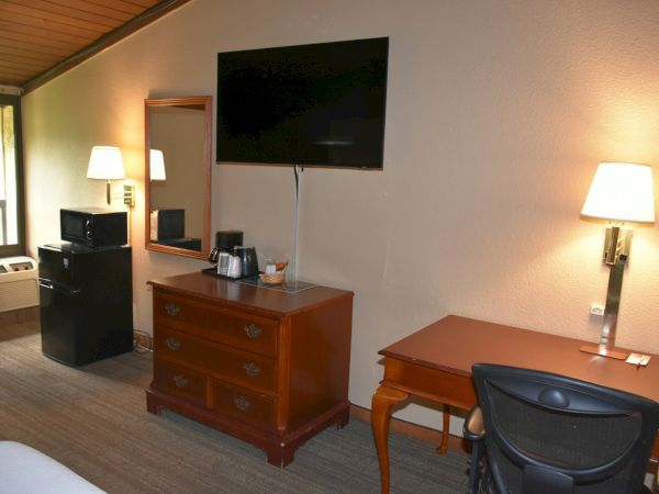 A hotel room with a desk, chair, wall-mounted TV, dresser, mini-fridge, coffee setup, and a window with a seating area, under warm lighting.