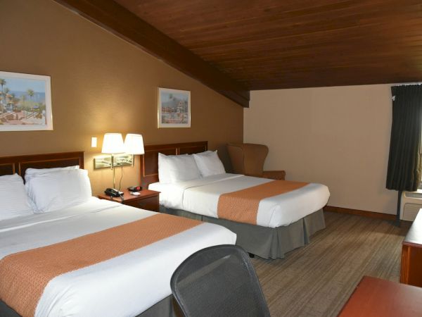 A cozy hotel room with two large beds, warm lighting, framed art, a sofa, wooden desk, and a window near the corner, inviting and neat.