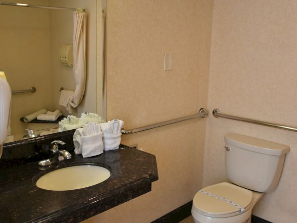 A small hotel bathroom with a dark countertop sink, mirror, exposed plumbing, toilet, beige walls, and a grab bar along the wall.