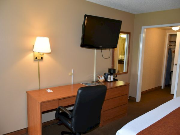 A hotel room with a desk, chair, wall lamp, mounted TV, coffee maker, and a bed edge visible, plus a doorway to a bathroom.