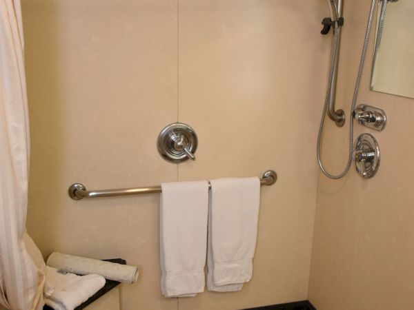 A beige tiled bathroom with a curved shower rod, overhead shower, wall grab bar, a towel bar with two white towels, a bench, and a small square floor drain.