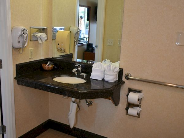 A hotel bathroom with a sink, mirror, countertop, stacked towels, soap dispensers, a toilet, grab bar, and a mirror reflection in the doorway.