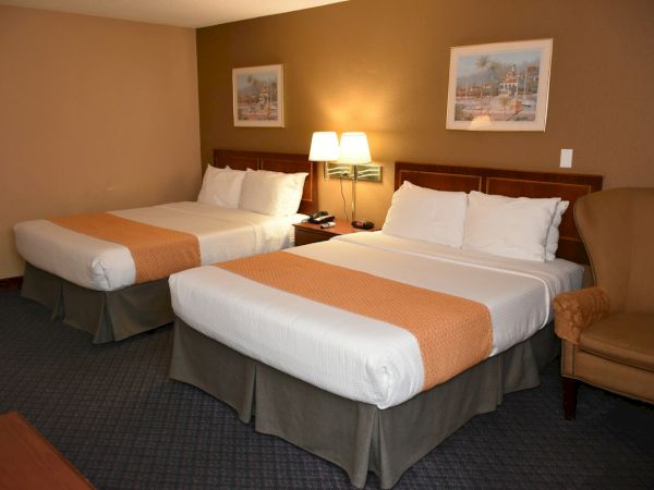 Two double beds in a hotel room with a nightstand, lamp, and a beige armchair, plus framed art on the wall, under warm lighting.