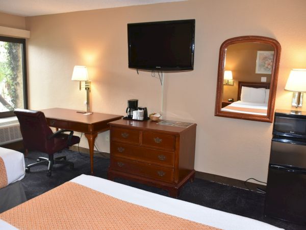 A hotel room with two beds, a desk and chair, wall-mounted TV, dresser, mirror, lamps, and a mini-fridge area.