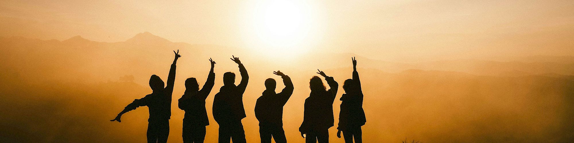 A group of people stands on a hill at sunset, silhouetted against a hazy orange sky with their arms raised in celebration.