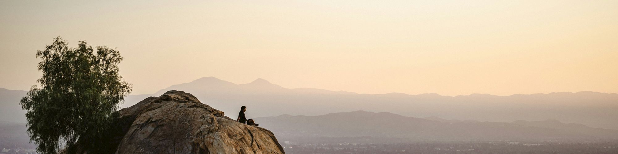 A person sits atop a large rocky outcrop overlooking a vast valley at sunset, with distant hills and a calm, hazy horizon.