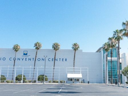 A large white convention center with "Ontario Convention Center" sign, palm trees lining the front, and a spacious empty parking lot under a clear blue sky.