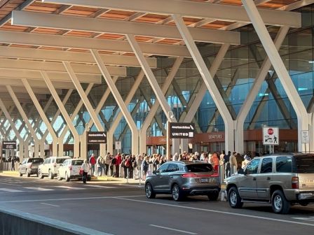 People waiting in line outside a modern airport terminal with white structural beams and glass, cars parked nearby, signage visible, sunny day.