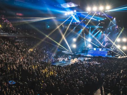 A massive concert with a packed crowd, bright stage lights and performers on a raised platform under a canopy of colorful beams.