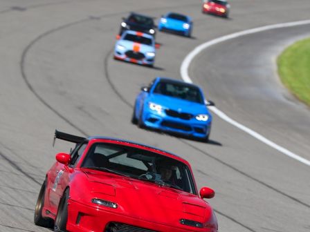 A red sports car leads a line of colorful race cars on a curving track, blue cars behind, with motion blur and grassy infield.