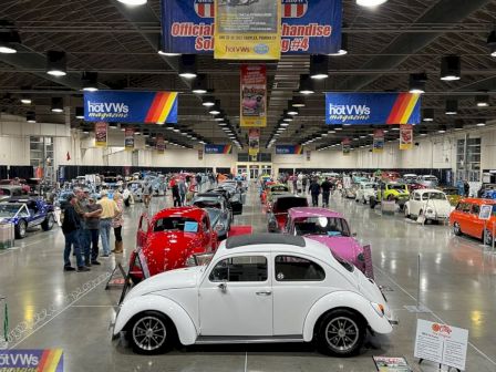 A spacious car show with classic VW Beetles in white, red, pink, and orange, people browsing, banners overhead, and polished exhibition space.