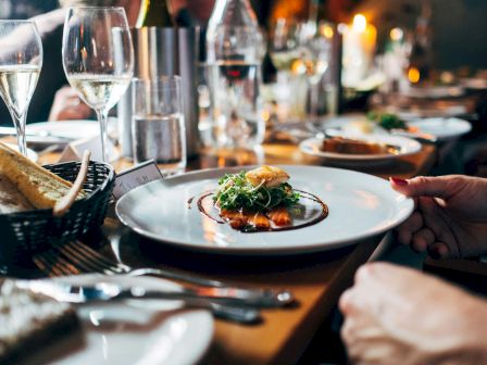 A fancy plated dish at a dinner table with wine glasses, bread basket, and hands ready to plate; fine dining ambiance.