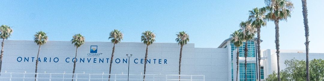 A large white convention center building labeled &ldquo;Ontario Convention Center&rdquo; with palm trees lining the front, a clear blue sky, and an empty parking lot.