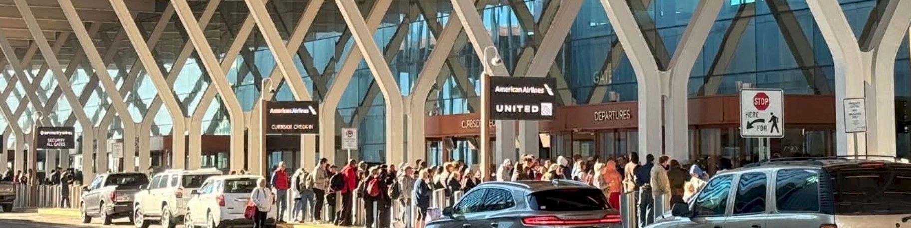 A queue of people at an airport curb under a modern, lattice-like roof, with cars lined up and signage visible near the terminal entrance.