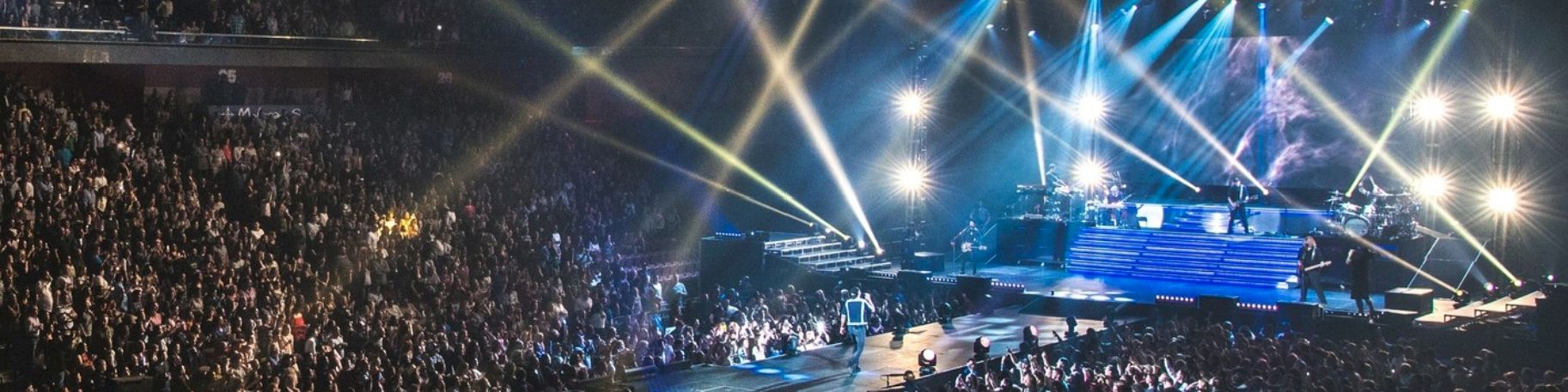 A packed concert arena with a massive crowd, illuminated stage lights, and a performer at center stage under bright beams, energizing the audience.